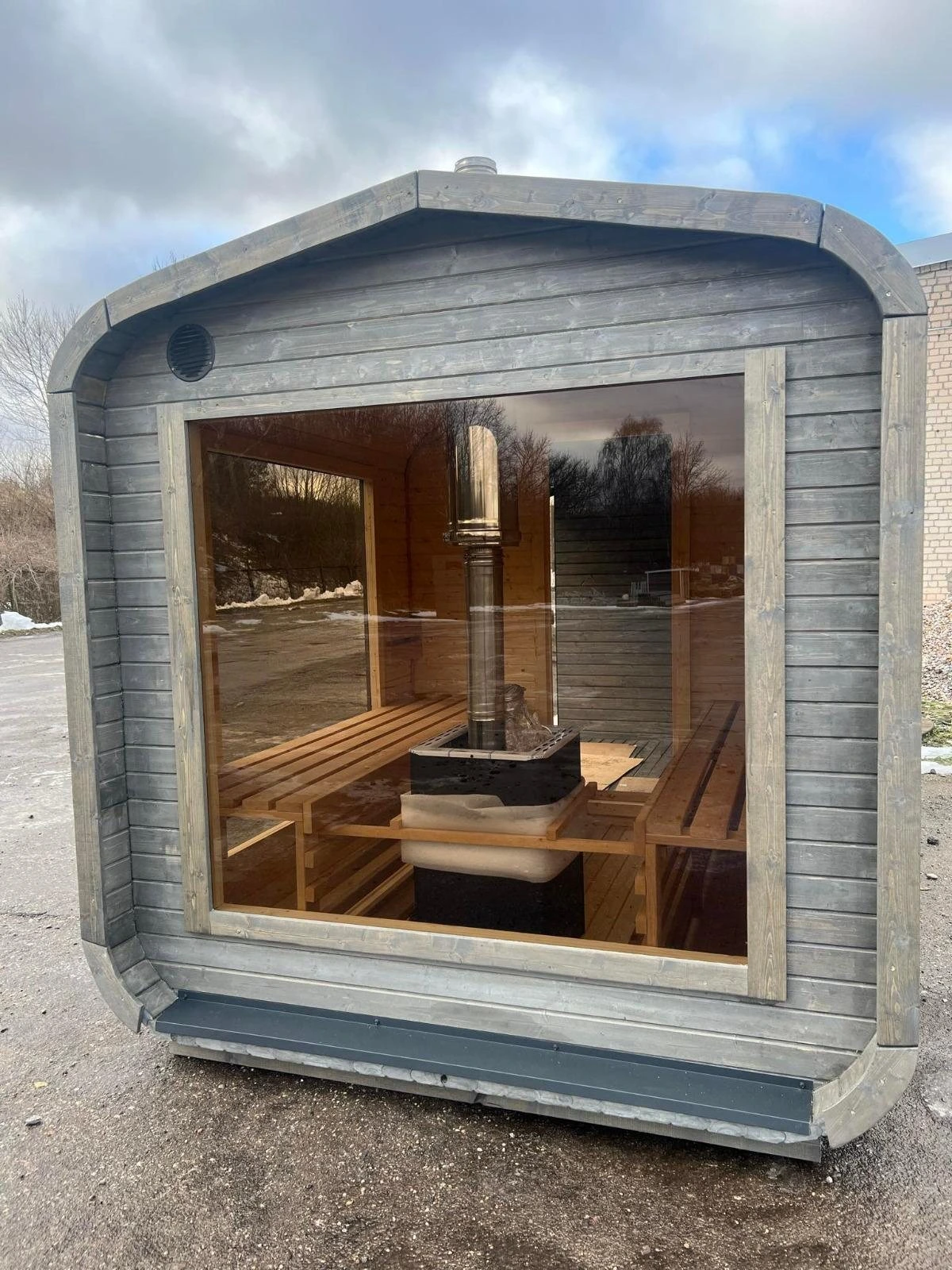 Grå badstue med avrundede hjørner og stort panoramavindu som viser benker og ovn
